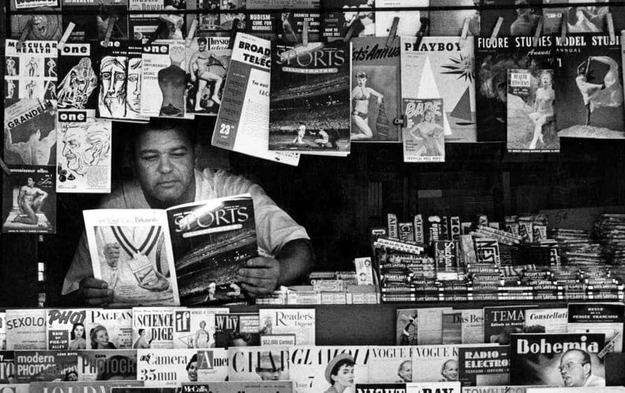 At a newsstand, a vendor reads an issue of Sports Illustrated magazine as he waits for customers, New York, New York, August 1954.