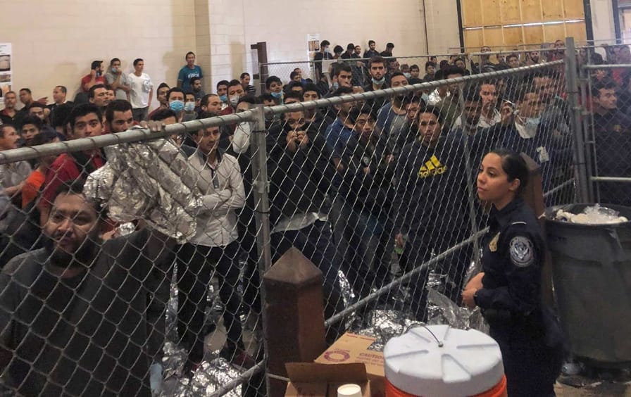 A crowded detention center with migrants looking in from behind a fence.