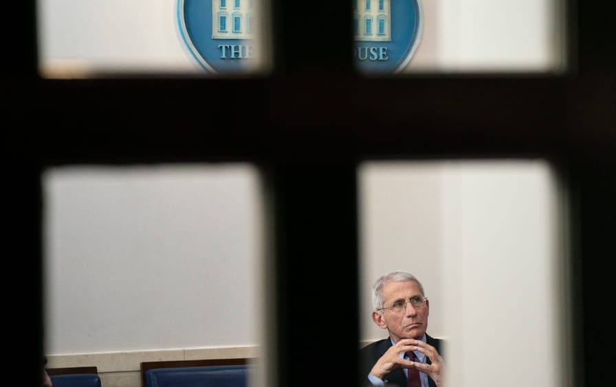 Dr. Anthony Fauci, pictured through a window, listens to a press briefing.