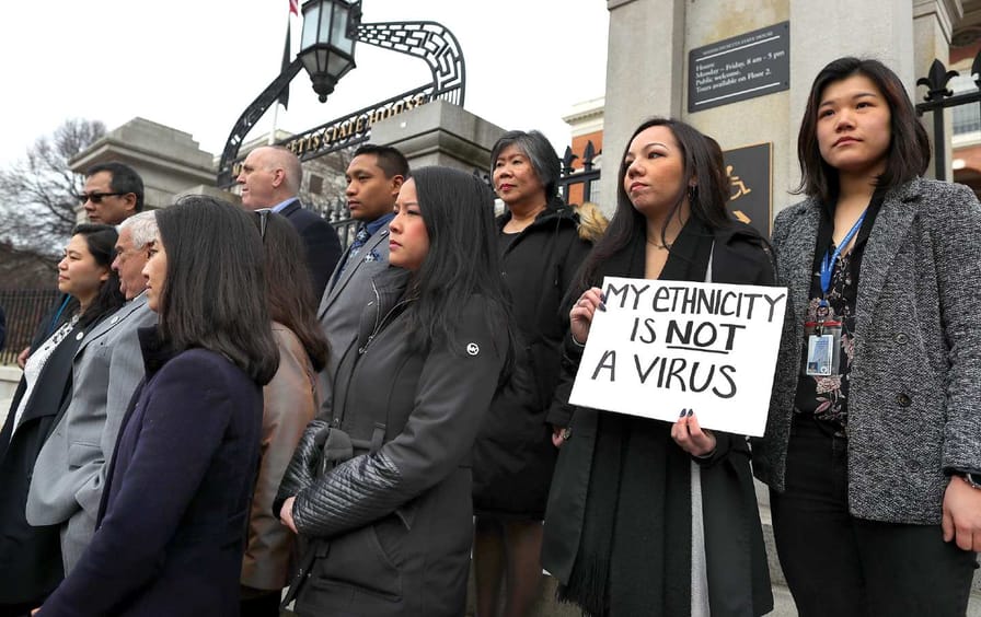 Asian Americans stand together on the steps of a building, one holds a sign reading 