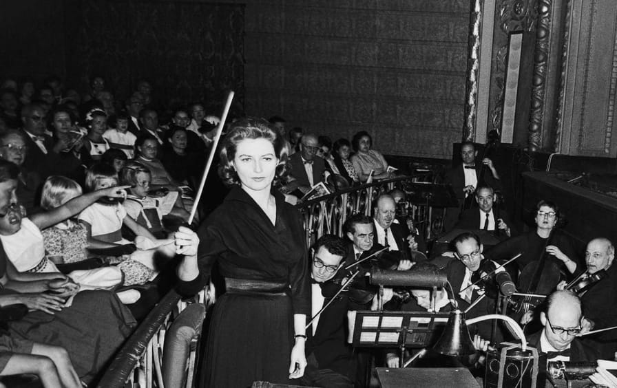 A conductor raises her baton in an orchestra pit, surrounded by musicians