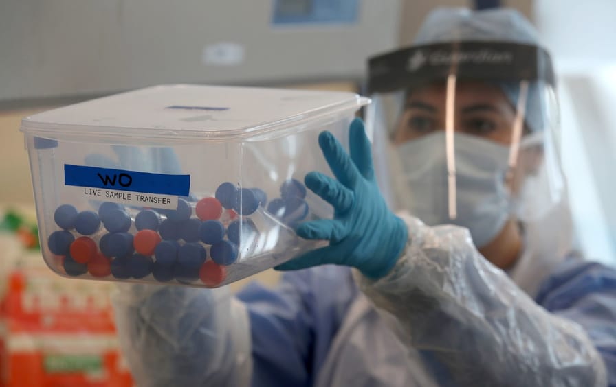 A lab technician in full protective gear holds a container of test tubes