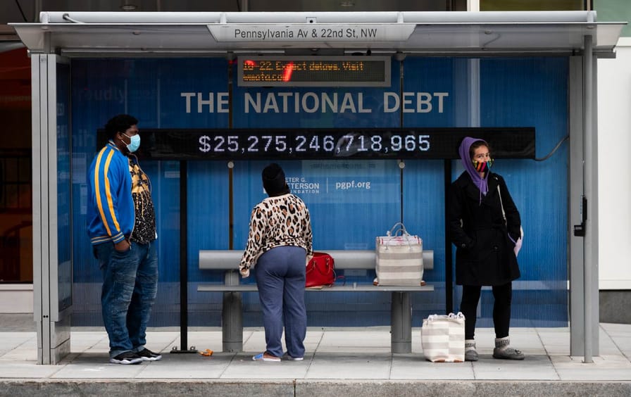 Passengers wearing face masks wait for their bus in front of a national debt display