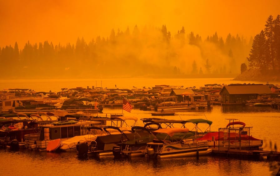 The sky is orange and smoky above the lake, where several boats are docked.