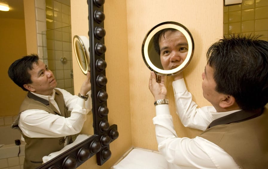 A man in a housekeeping uniform adjusts a mirror in a bathroom.