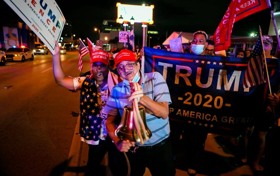 Two supporters in red MAGA hats pose for the camera, behind them is a supporter holding a Trump 2020 sign.