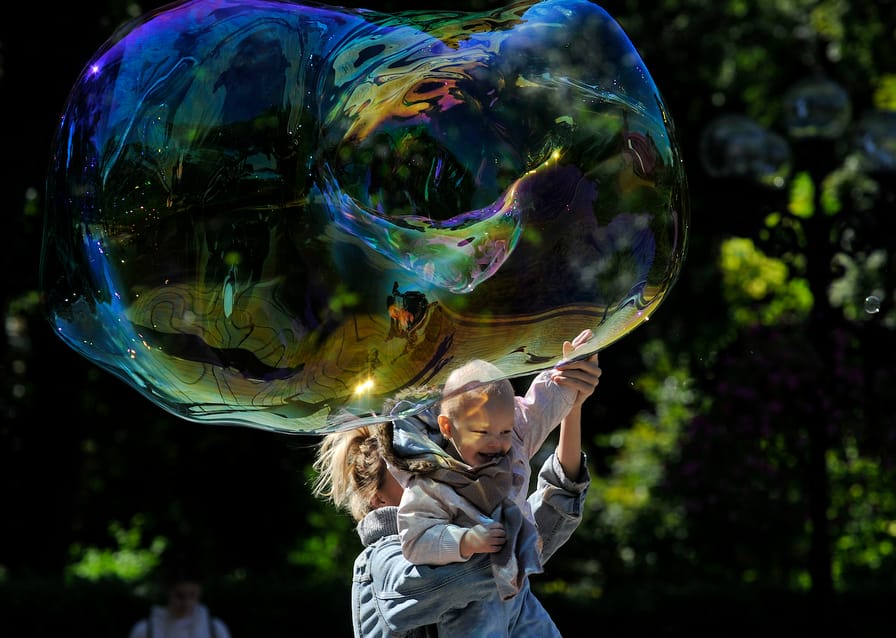 [Photo: A woman carries a child to get him to touch a huge soap bubble in the park.]