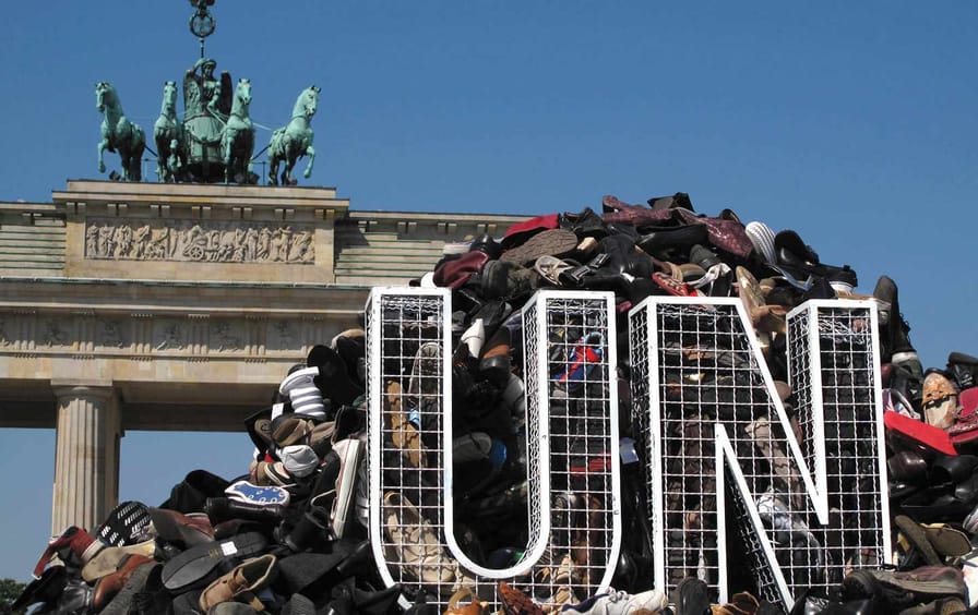 Wireframe sculptures of the letters U and N are placed in front of a large pile of shoes.