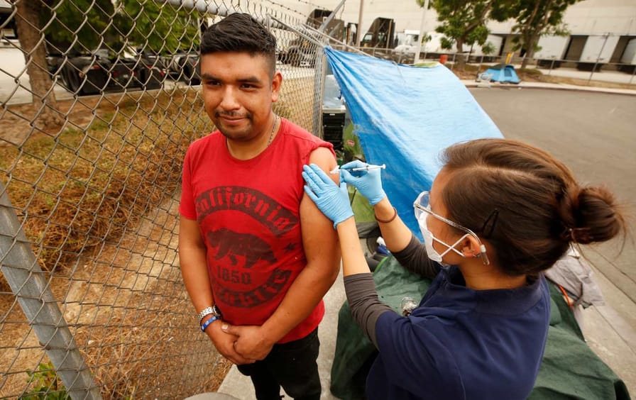 A man in a sleeveless red shirt receives a Covid vaccine in front of a tarp.
