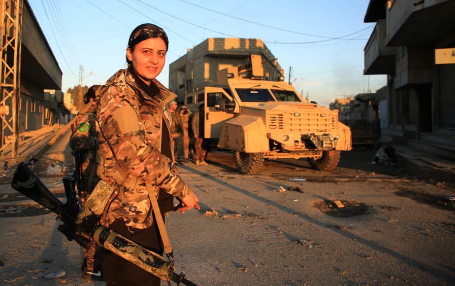 A female soldier looks toward camera, carrying a gun strapped to her shoulder. Other soldiers behind her congregate around an armored vehicle.