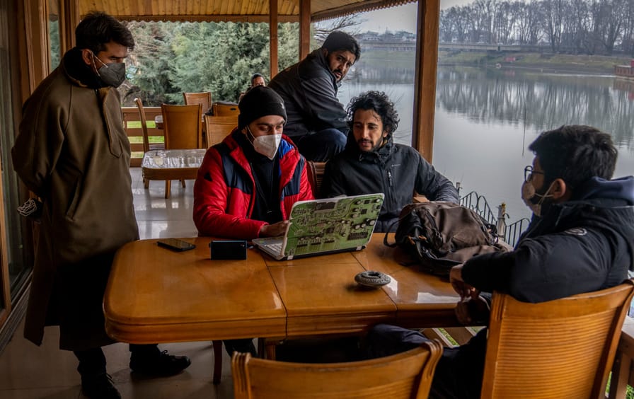 Five men sit around a table, where one is at work on a laptop