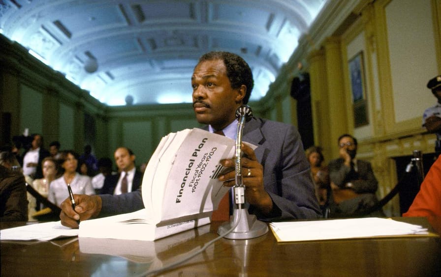 A Black man in a suit sits at a table with a thick booklet in front of a crowd.