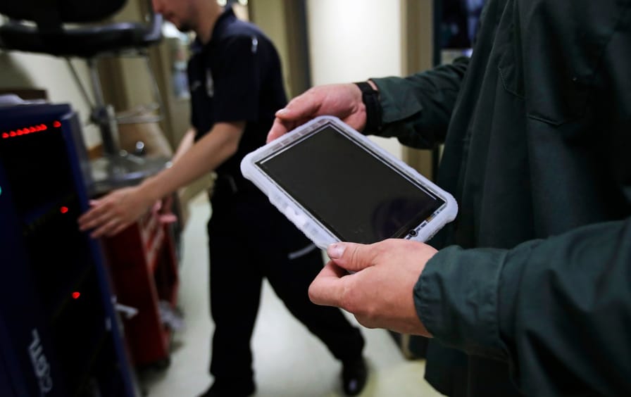 A man's hands hold a prison tablet while another man walks in the background