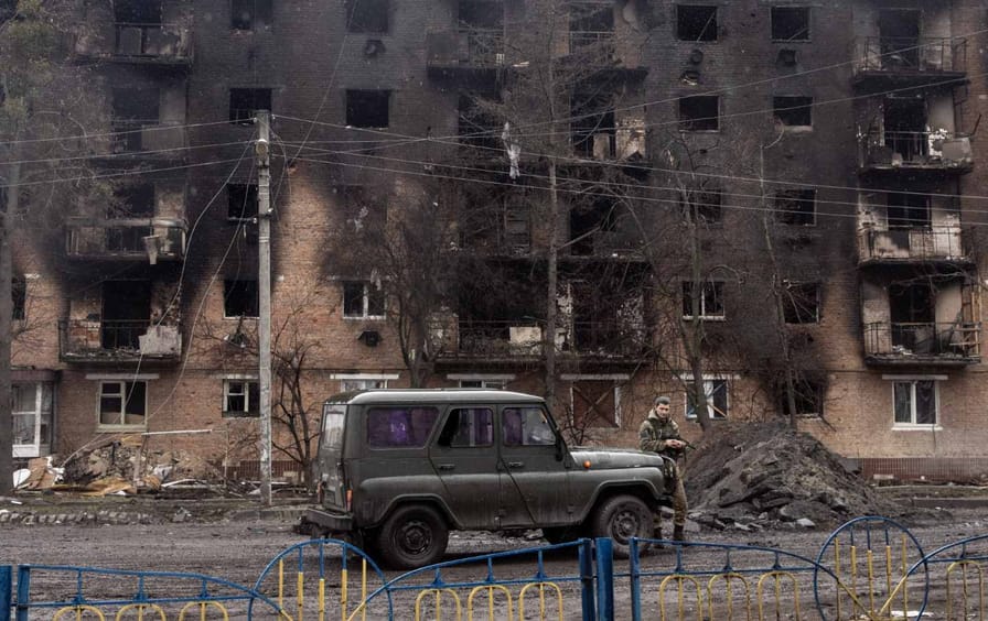 A car sits in front of a burned apartment building and pile of rubble. A soldier stands in front of the car.