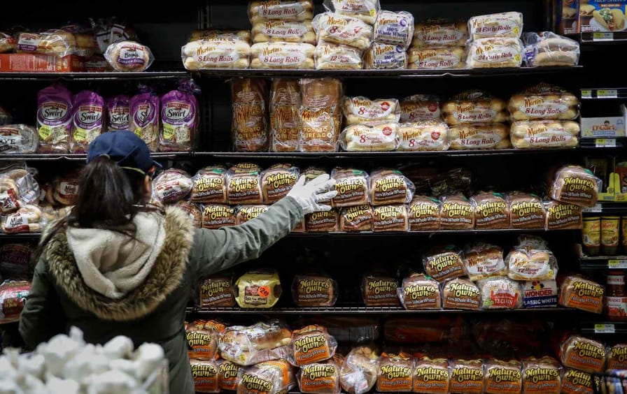 A woman wearing gloves picks out a loaf of bread at the supermarket