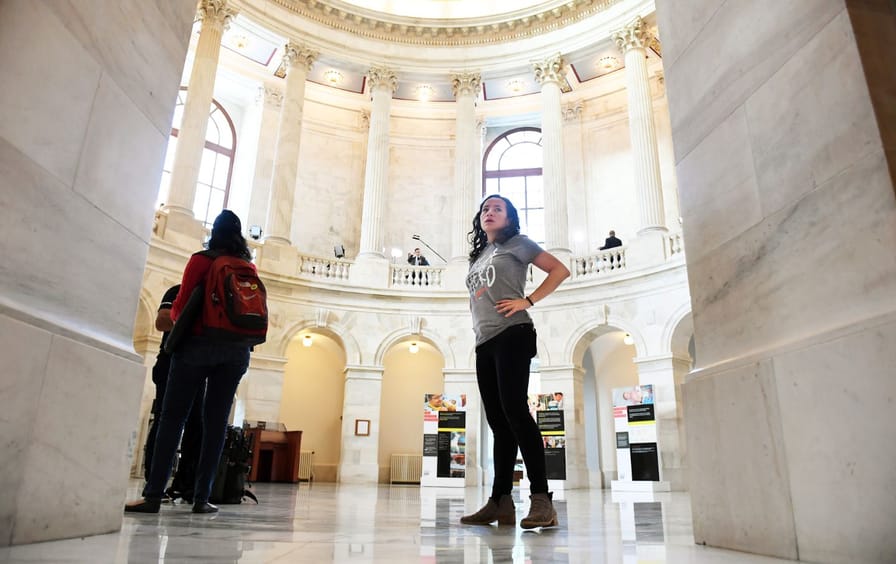 A woman in a gray T-shirt and black jeans stands in a white room with pillars.
