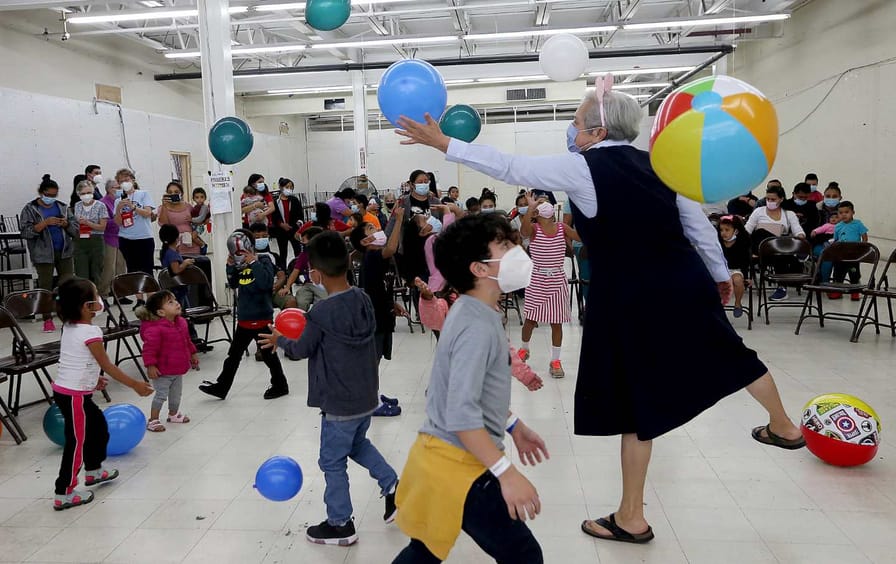 Masked children, led by a nun, play with beach balls and balloons while adults watch.