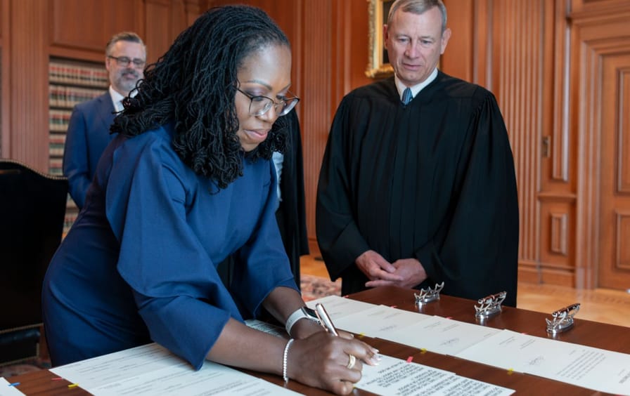 A woman signs a document as a man in judicial robes watches.