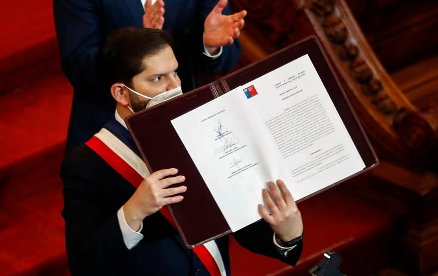 A man in a Covid mask, red, white and blue sash and black suit holds up a signed document.