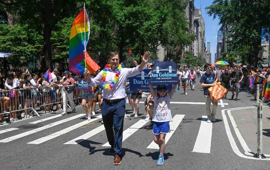 Dan Goldman marching at NYC Pride with his daughter. He's holding a pride flag and wearing a rainbow lei. His daughter holds a sign reading 