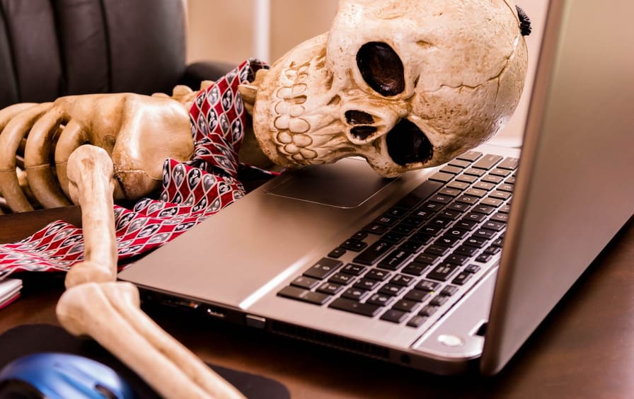 A skeleton worker with a tie on lays its head on the computer, exhausted.