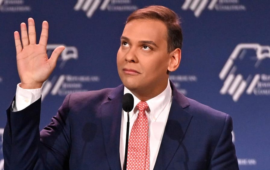 Headshot of George Santos speaks in Las Vegas at the Republican Jewish Coalition (RJC) annual meeting. His hand is raised in oath.