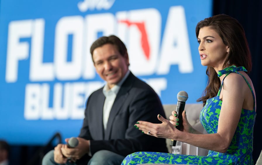 Casey DeSantis, in the foreground, speaks holding a microphone while Ron DeSantis looks on. They are in front of a large banner reading 