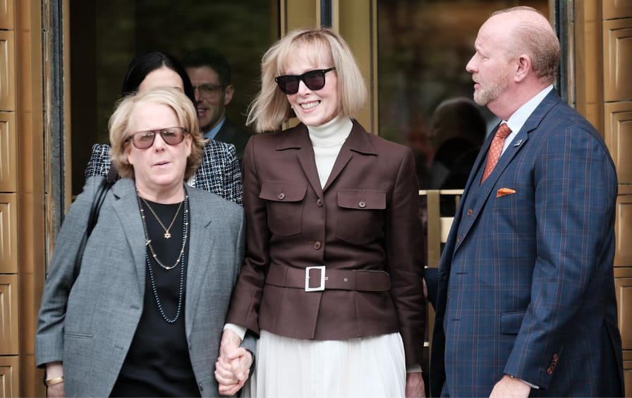 E. Jean Carroll, smiling, holds hands with her attorney, Roberta Kaplan, at her side, as they leave a court house.