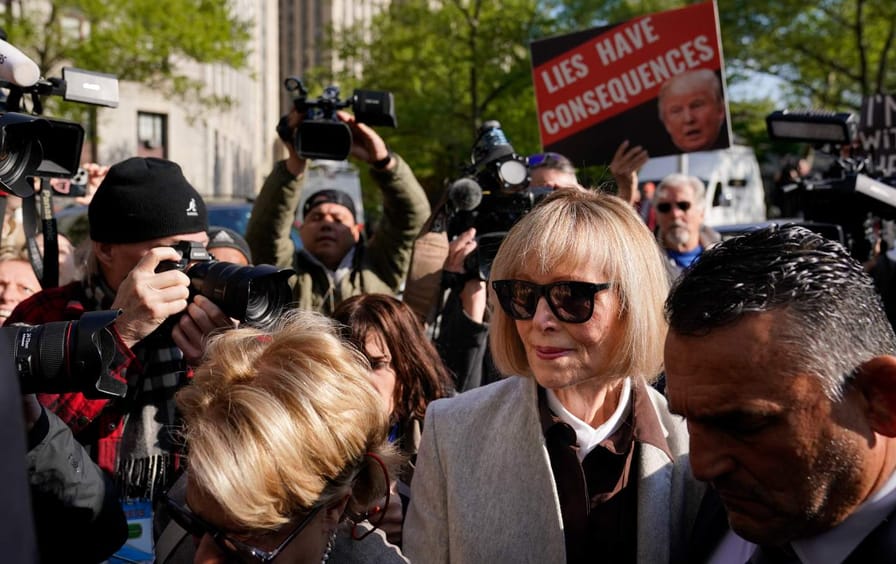 E. Jean Carroll outside Manhattan federal court house surrounded by a scrum of people, one holding a sign saying 