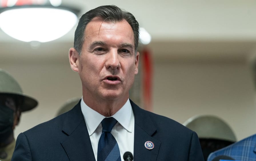 A photo of Tom Suozzi, wearing a coat and tie, speaking indoors.