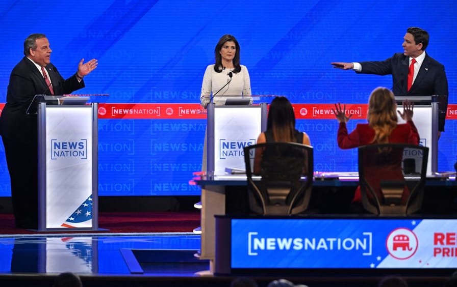 On the debate stage, Chris Christie and Ron DeSantis gesture and point toward each other while Nikki Haley looks on between them.