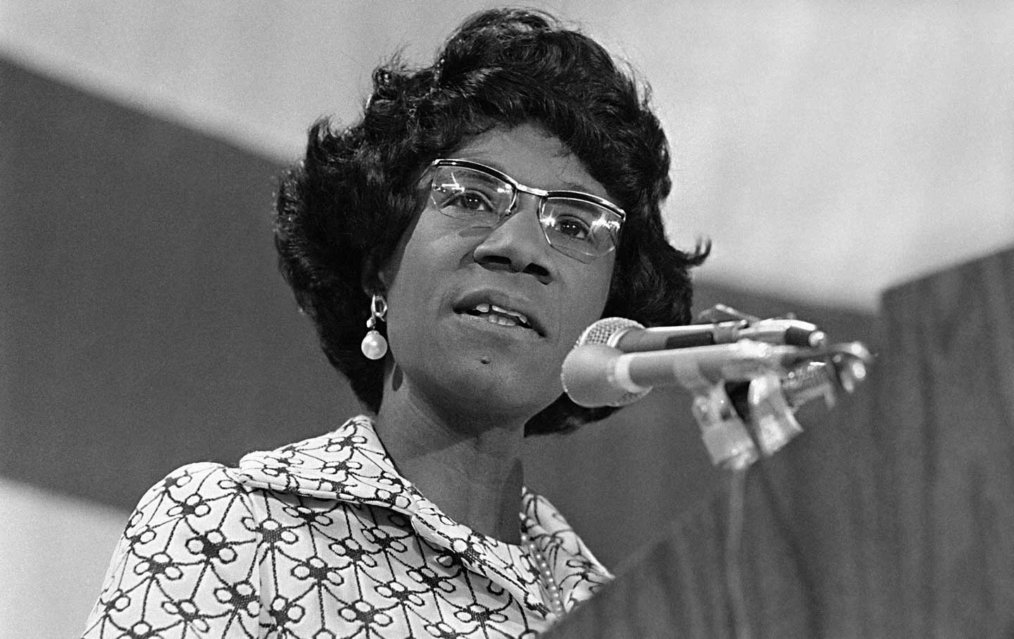 This black-and-white photo of Shirley Chisholm shows her standing on a dais while speaking into a microphone.