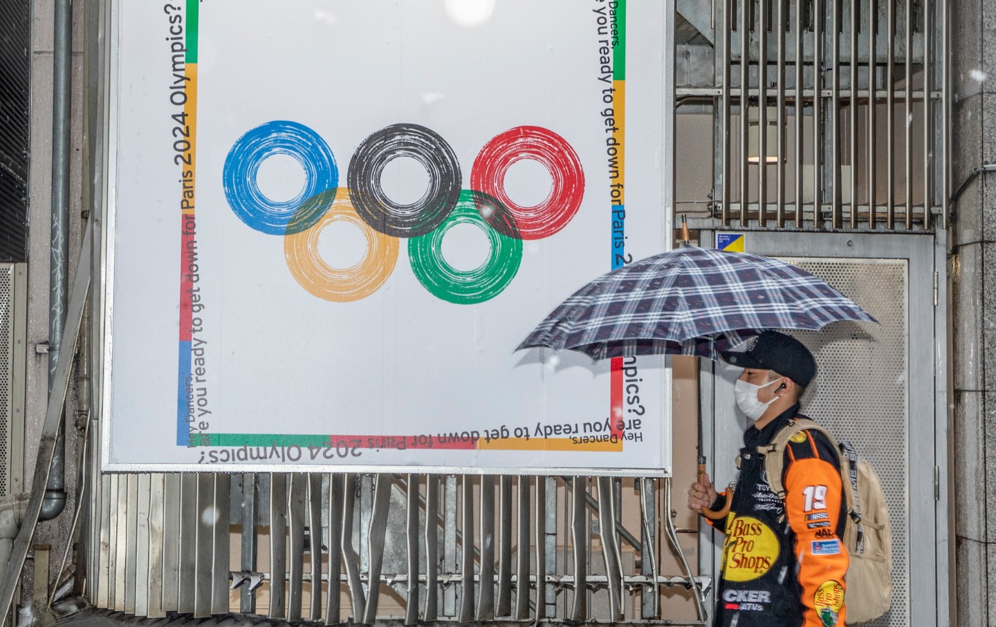 A man in a mask walks by an Olympic 2024 poster