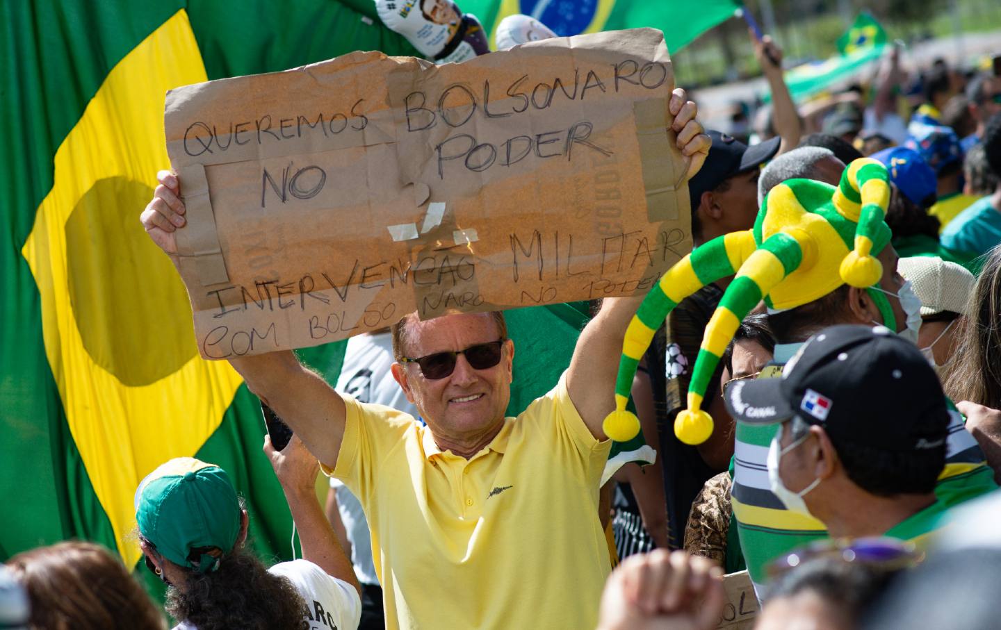 A man with sunglasses holds a cardboard sign, with writing in Portuguese.
