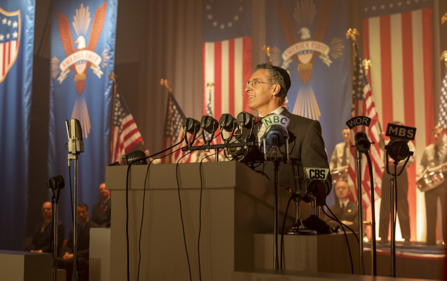 John Turturro stands at a podium with microphones, playing a rabbi in The Plot Against America