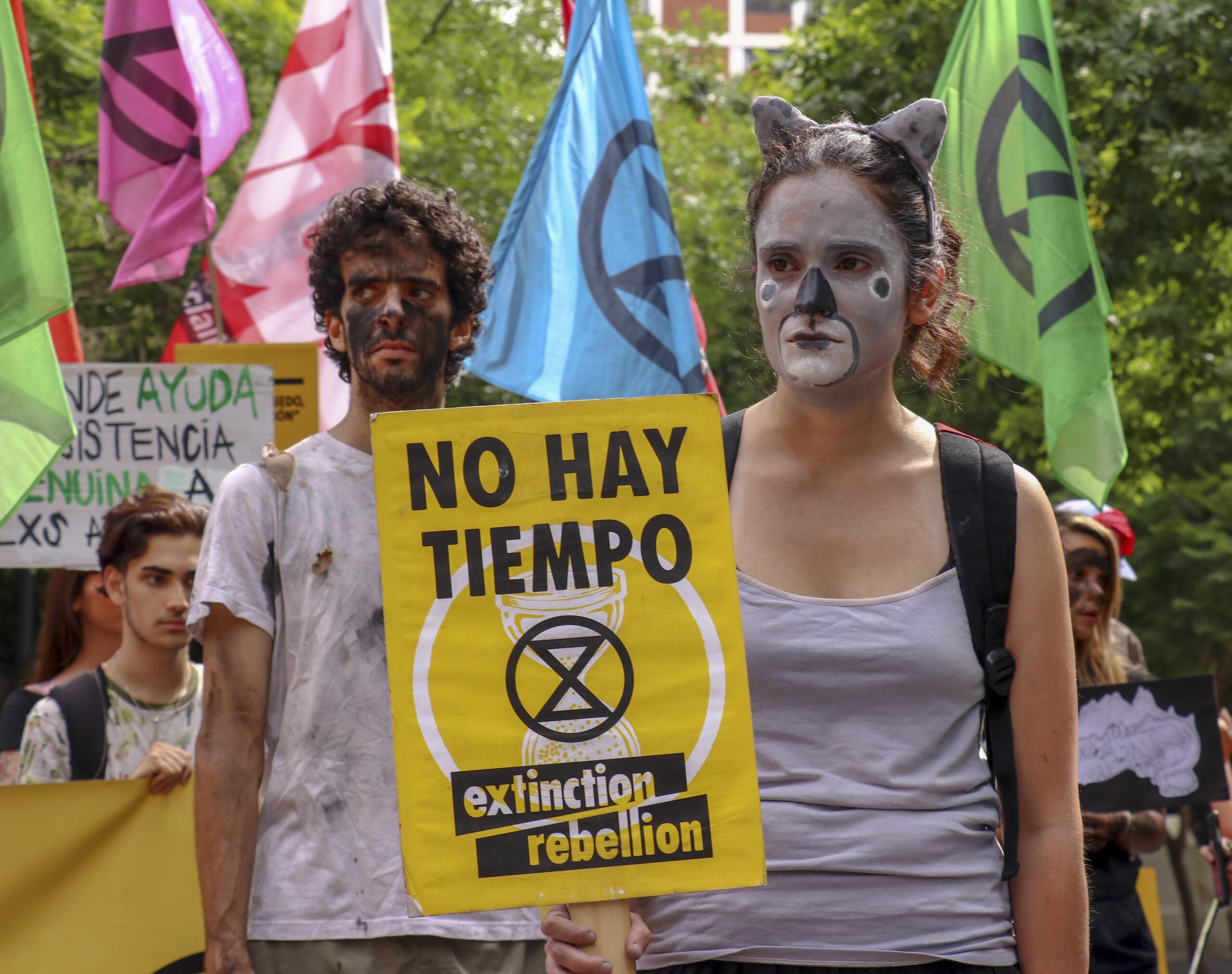 Two activists with painted faces stand, holding a sign that reads 