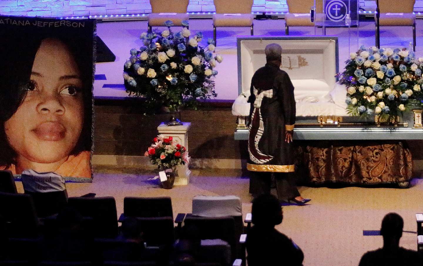 A woman dressed in black stands over Atatiana Jefferson's casket, while others wait behind her. A large photo of Jefferson is displayed to the left of the casket