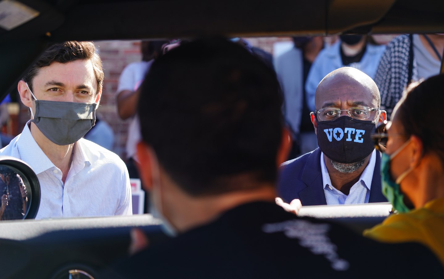 Jon Ossoff and Raphael Warnock, both wearing masks, speak to supporters who are seated in their car.