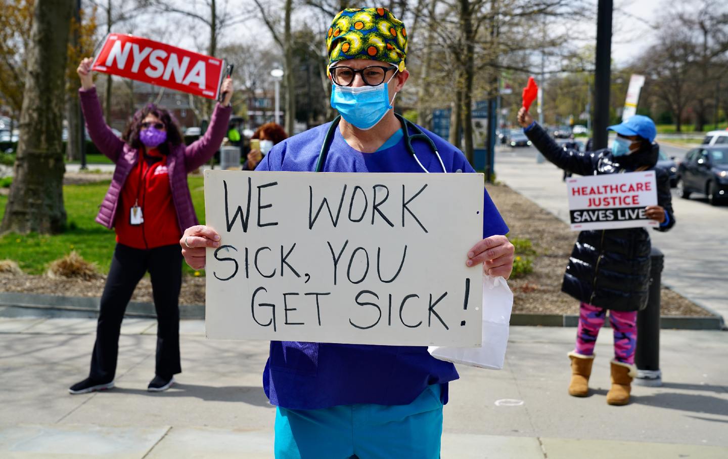 Three people protest, holding signs and wearing masks. The one in the center front holds a sign that reads 