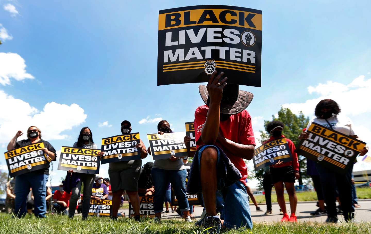 A man in a hat kneels while holding a sign that says 