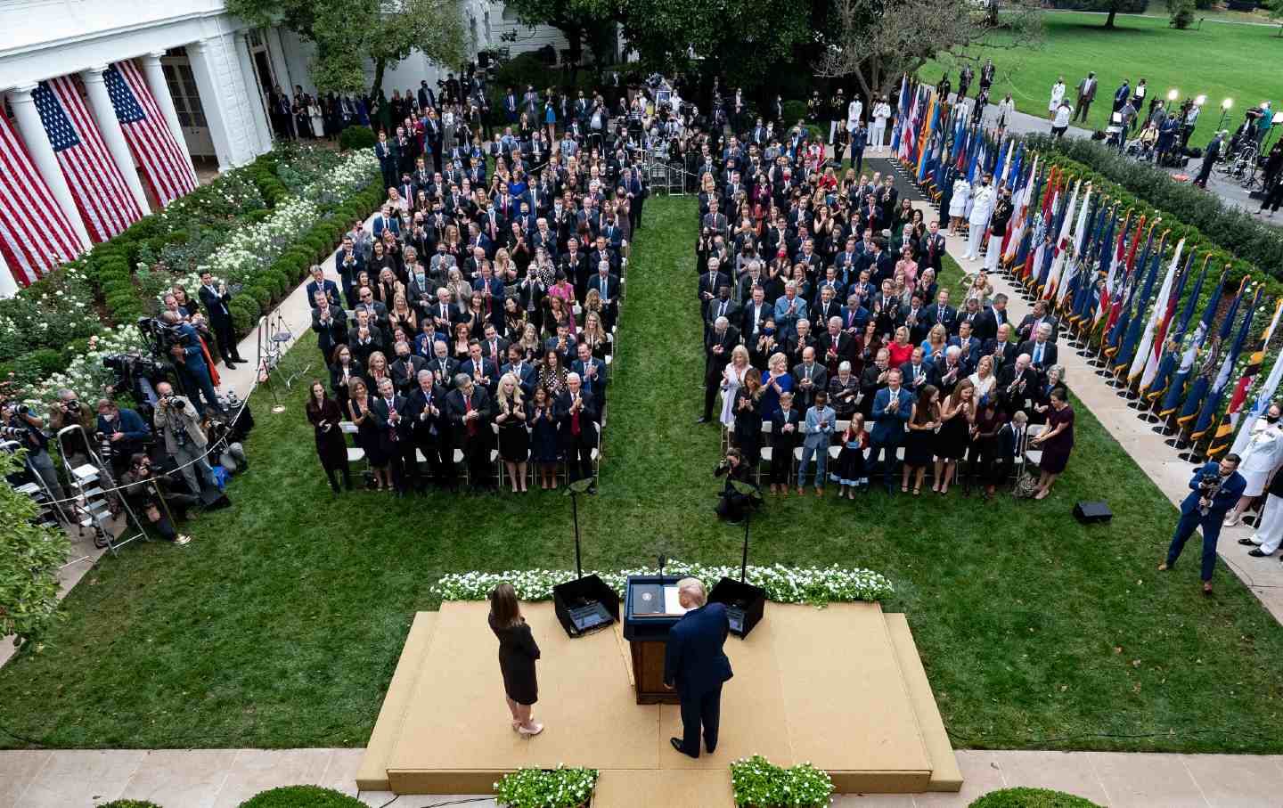 Amy Coney Barrett and Donald Trump stand on a platform facing a crowd at an outdoor press conference.