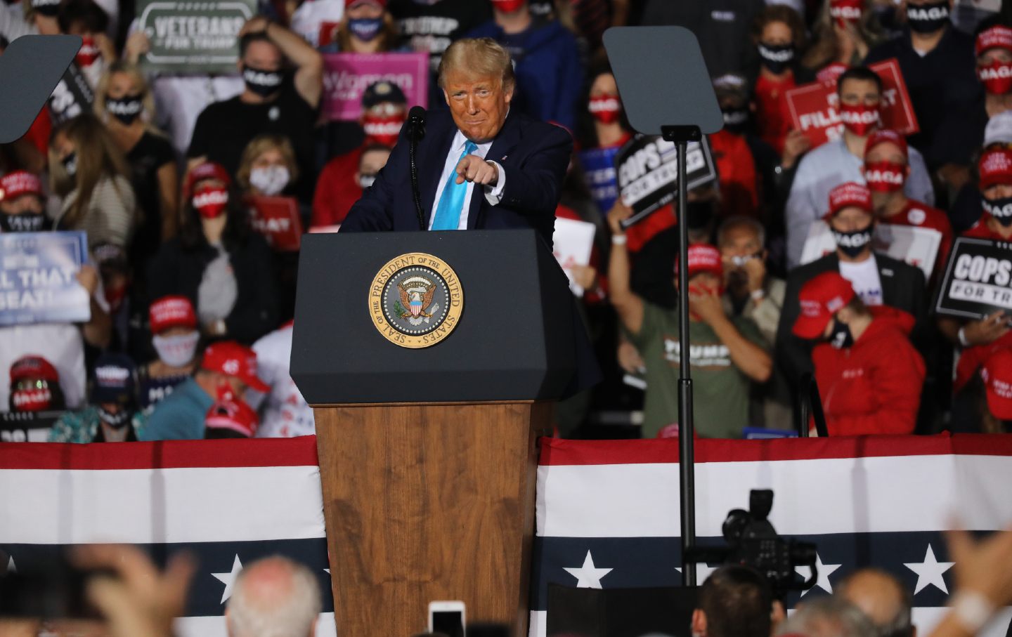 Donald Trump points at the crowd from the stage at a rally as people behind him wave signs supporting him.