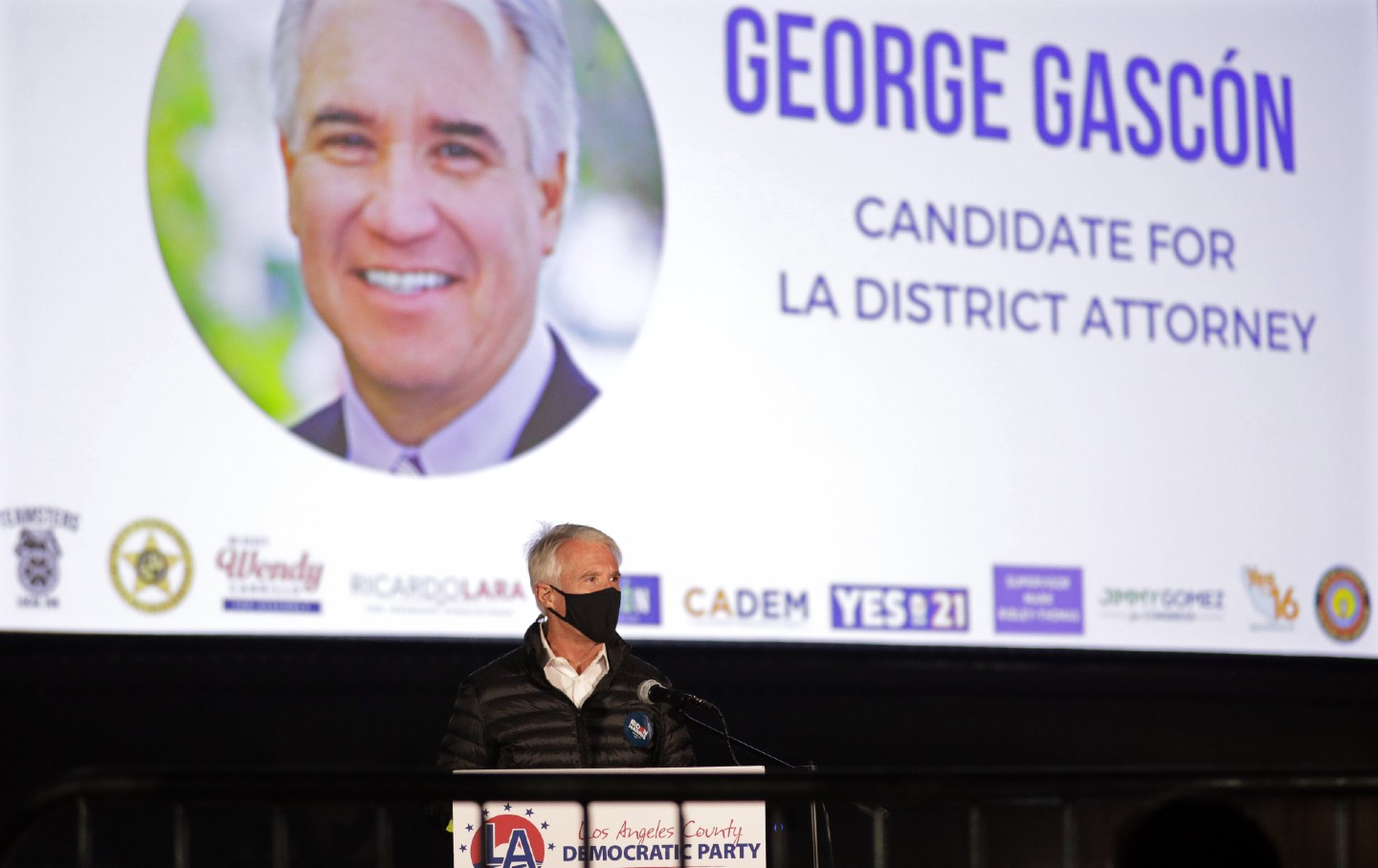 Gascón stands, wearing a mask, in front of a large projected slide with a picture of himself that reads 