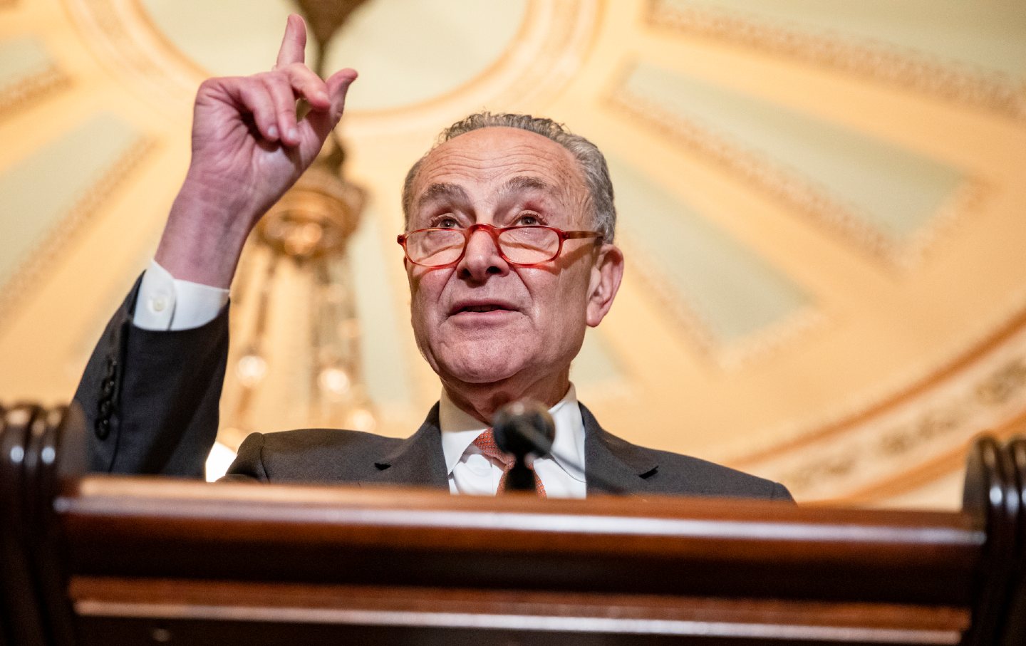 Chuck Schumer stands behind a podium, wagging a finger. He wears a suit and tie and red glasses.