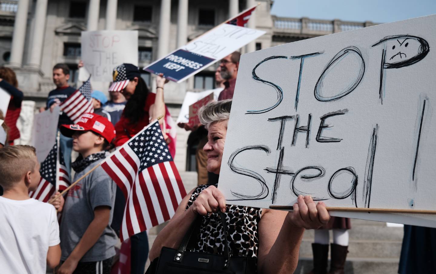 A crowd of white people adorned with American flags stand outside the Pennsylvania capitol, one sign says 