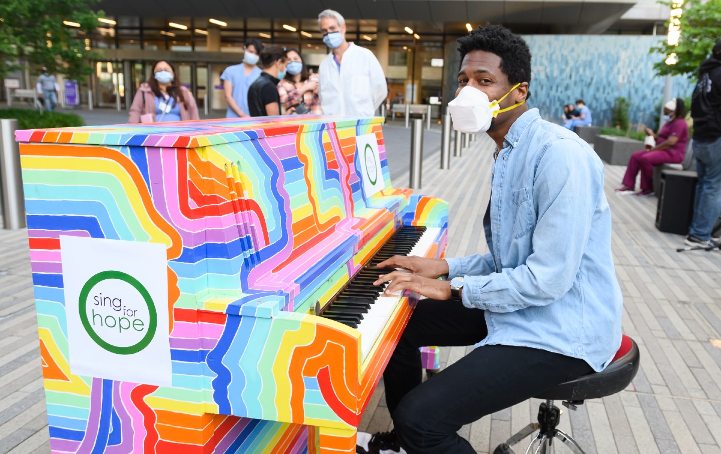 Jon Batiste plays a rainbow-colored piano outdoors as people in surgical masks walk by