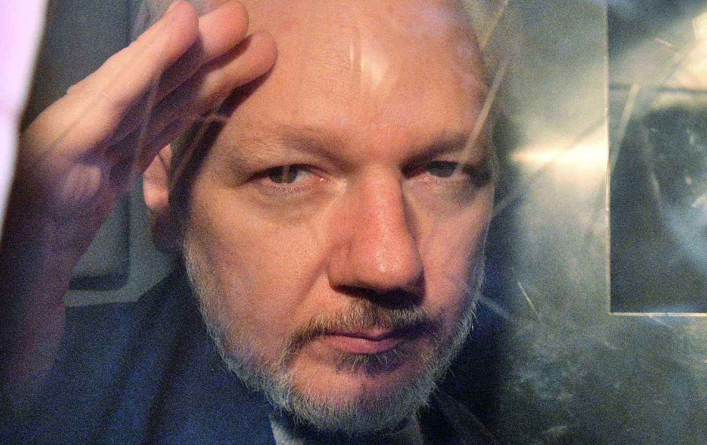Julian Assange waves slightly with his face up close against the window of a prison van