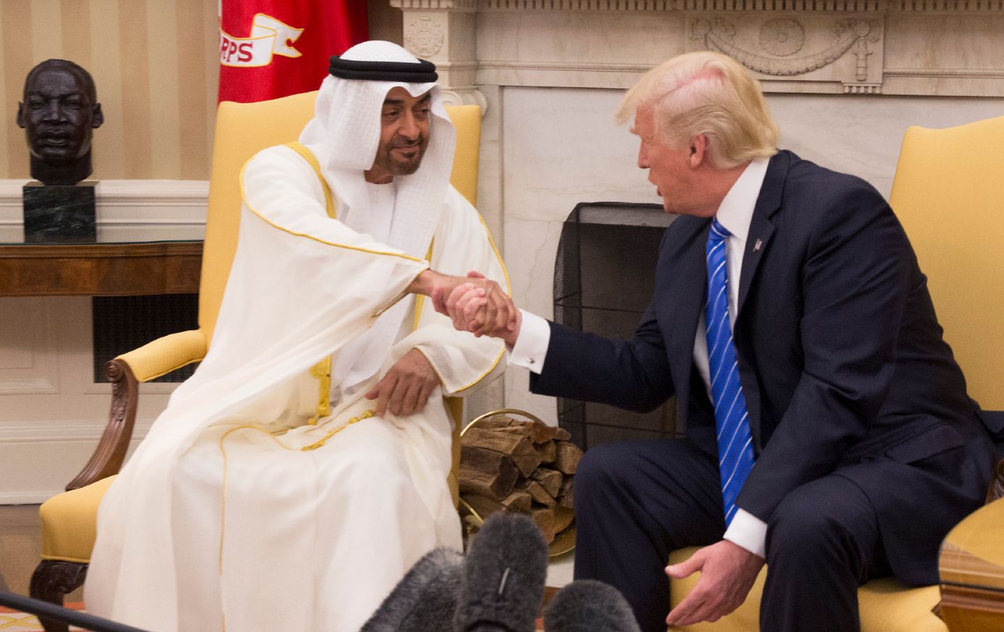 On the left, Shaikh Mohammed bin Zayed Al Nahyan sits and shakes Donald Trump's hand, on the right.
