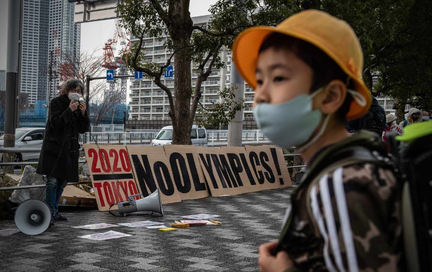 A young schoolboy in a mask, wearing a black jacket with three white stripes and an orange bucket hat, walks past a banner saying “No Olympics” during a protest calling for the Tokyo Olympics to be canceled.
