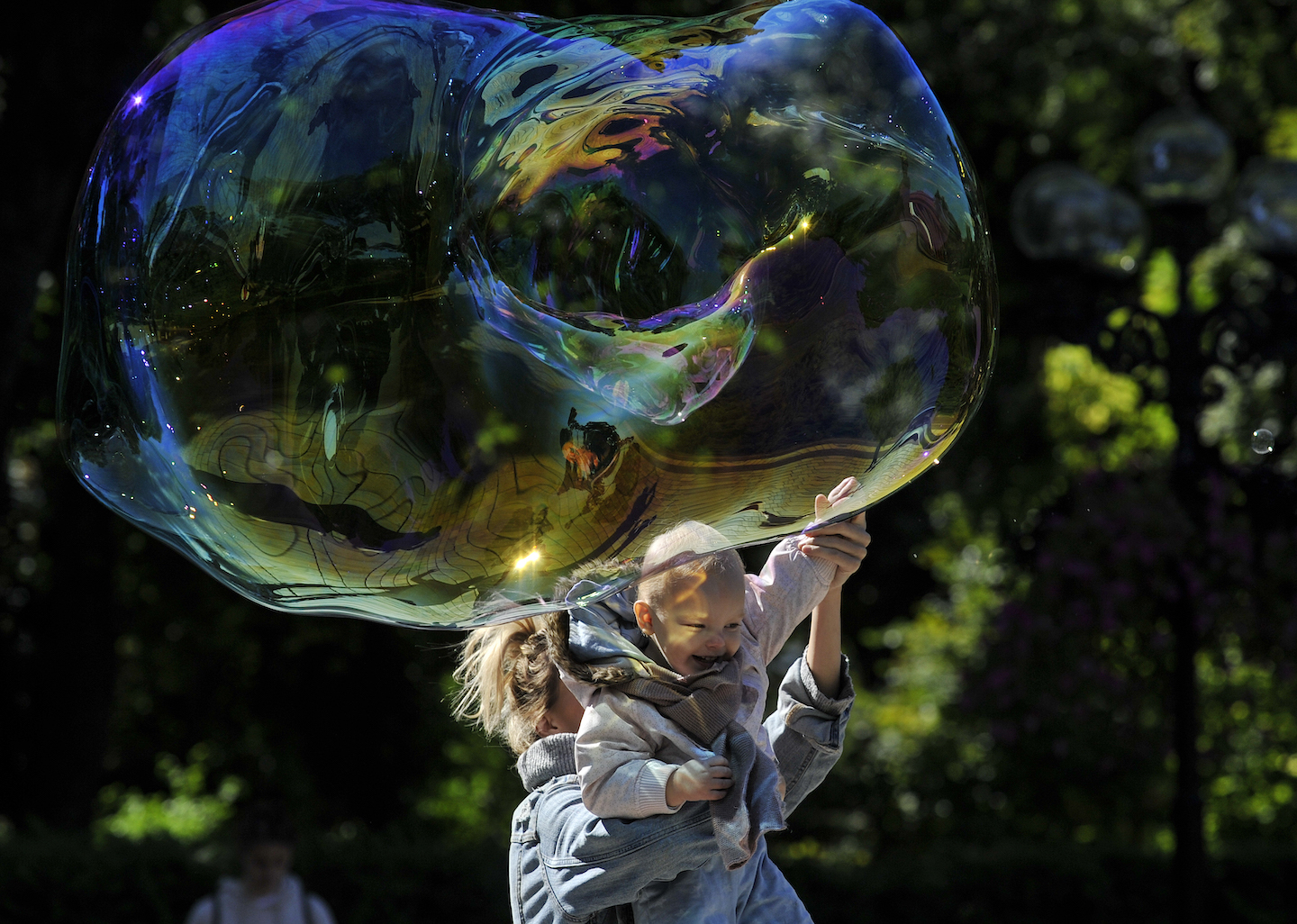 [Photo: A woman carries a child to get him to touch a huge soap bubble in the park.]